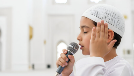 A young reciter calling the Adhan into a microphone, one hand cupped to his ear, inside a mosque